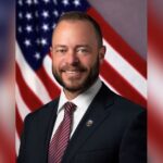 Tustin Mayor Austin Lumbard in a suit and City of Tustin lapel pin, smiling in front of an American flag