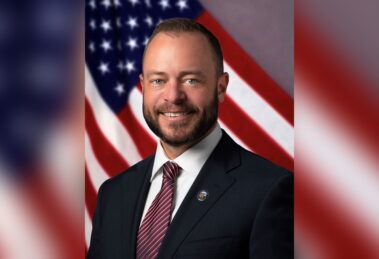Tustin Mayor Austin Lumbard in a suit and City of Tustin lapel pin, smiling in front of an American flag