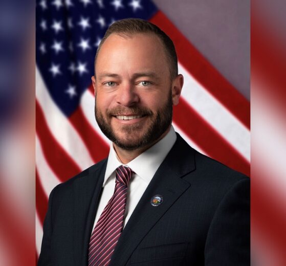 Tustin Mayor Austin Lumbard in a suit and City of Tustin lapel pin, smiling in front of an American flag