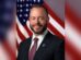 Tustin Mayor Austin Lumbard in a suit and City of Tustin lapel pin, smiling in front of an American flag