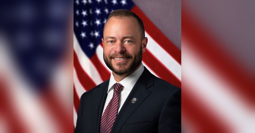 Tustin Mayor Austin Lumbard in a suit and City of Tustin lapel pin, smiling in front of an American flag